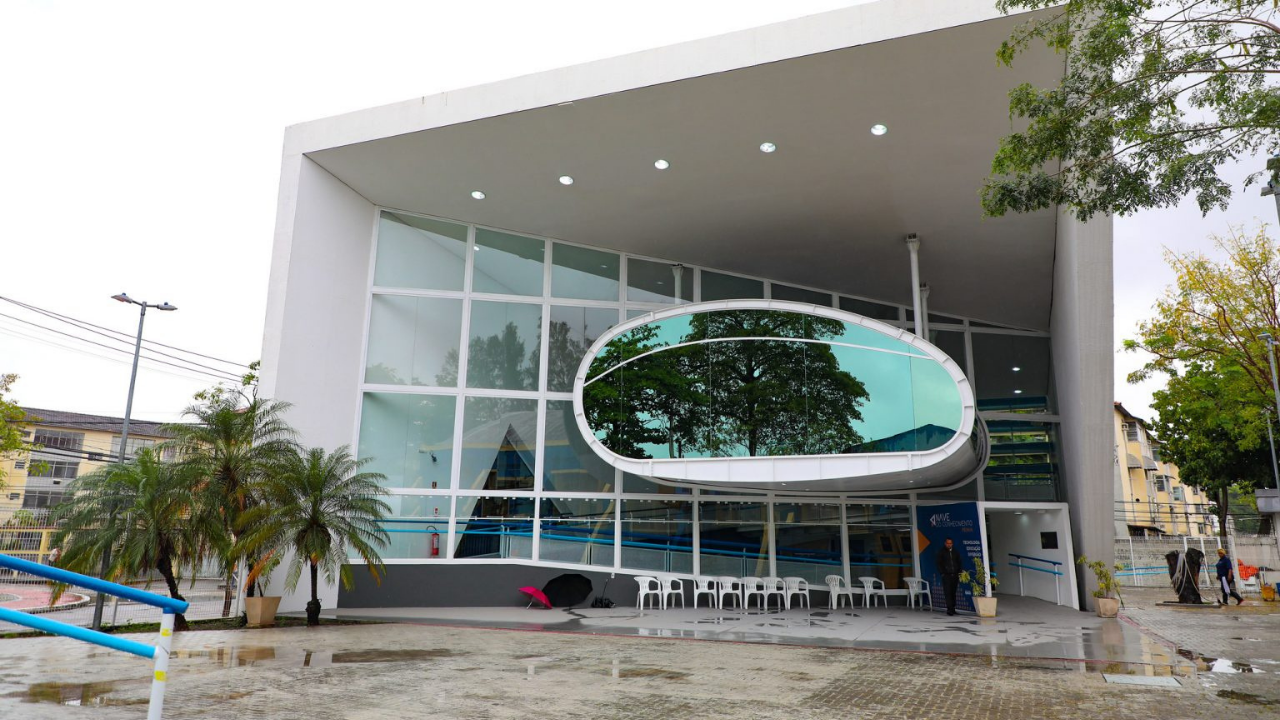 Fachada de uma unidade das Naves do Conhecimento com arquitetura moderna. O prédio possui estrutura branca, grandes painéis de vidro e uma abertura oval suspensa na fachada frontal. Em frente à entrada há cadeiras brancas alinhadas sob uma marquise. Árvores e vegetação aparecem nas laterais, e o chão molhado indica que a foto foi registrada após chuva. Algumas pessoas estão próximas à porta de acesso.