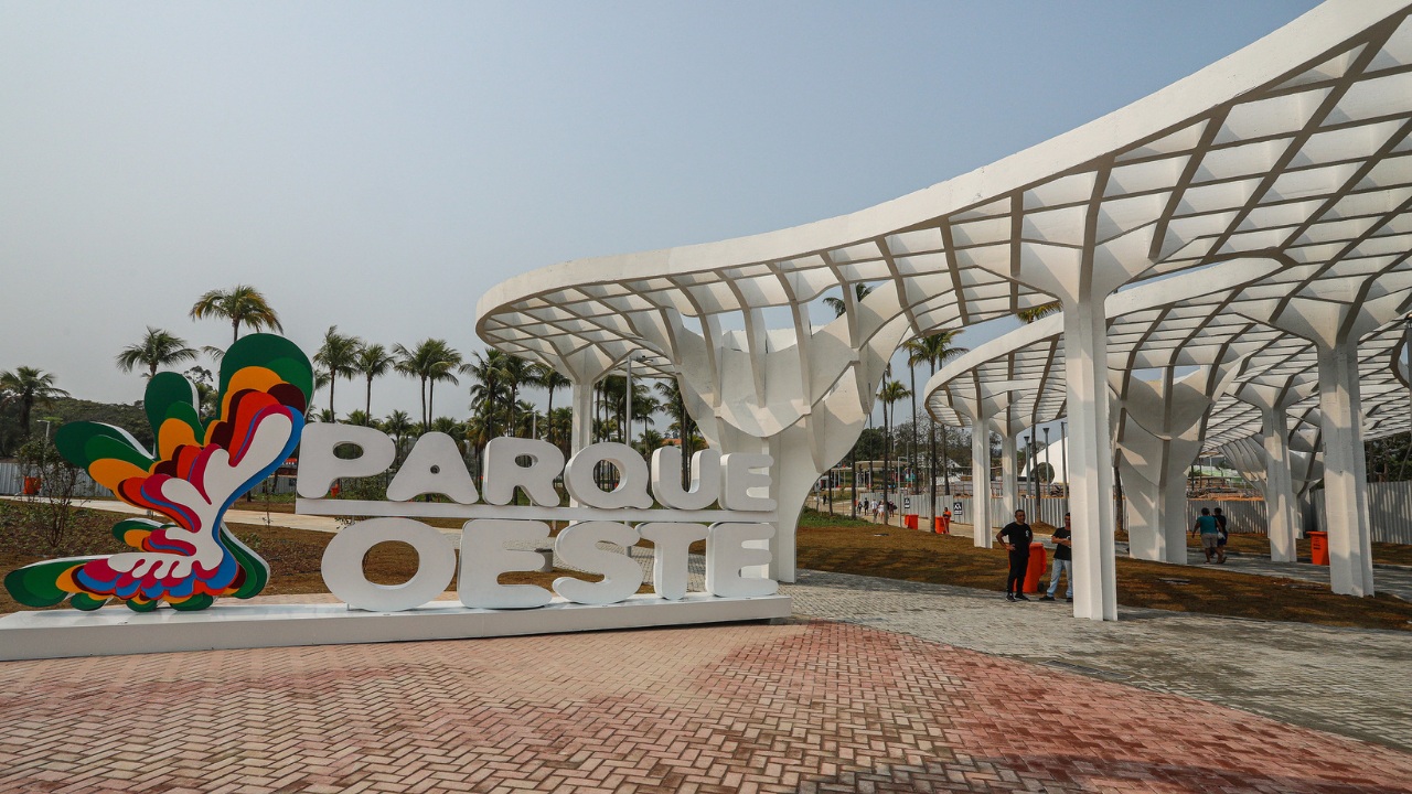 Entrada do Parque Oeste com grande letreiro branco escrito “Parque Oeste”, acompanhado de um símbolo colorido em formato orgânico com várias camadas nas cores verde, amarelo, laranja, vermelho e azul. O letreiro está instalado sobre piso de blocos intertravados. Ao fundo, estruturas arquitetônicas brancas em formato curvilíneo formam pérgolas que criam áreas de sombra ao longo do caminho. Há palmeiras distribuídas pelo espaço e algumas pessoas circulando pelo parque em um dia claro.