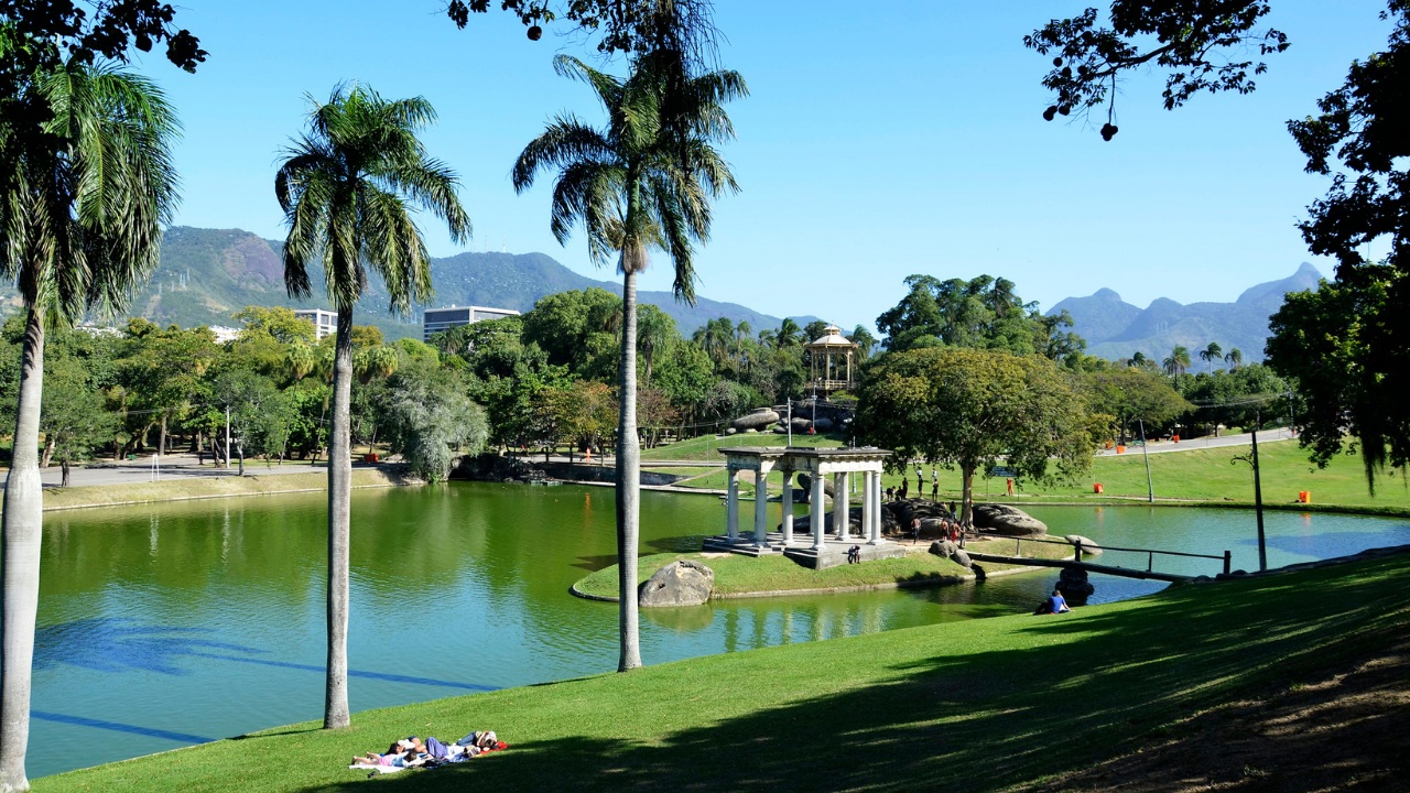 Paisagem ampla da Quinta da Boa Vista em um dia ensolarado. O lago de águas verdes ocupa o centro da imagem, cercado por gramados extensos e palmeiras altas. À direita, um pequeno conjunto de colunas brancas forma uma estrutura clássica sobre uma ilha do lago. Ao fundo, há muitas árvores, pessoas passeando e, mais distante, as montanhas do Rio de Janeiro recortam o horizonte.